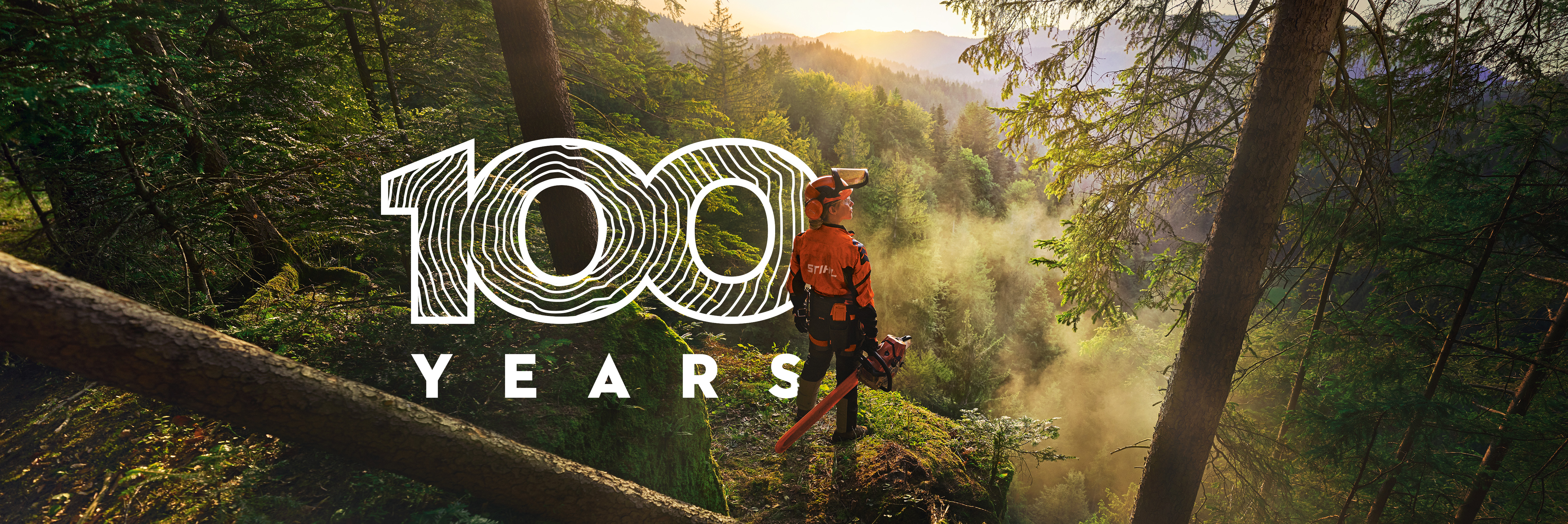Une femme se tient au bord d'une falaise dans une forêt d'un vert saturé. Elle porte des vêtements de travail STIHL orange, le casque ADVANCE ProCom et tient une tronçonneuse STIHL MS.