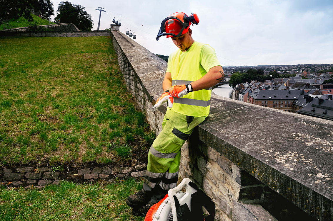 Jérémy s'appuie sur une clôture de la Citadelle de Namur avec un souffleur de feuilles et des gants de sécurité.