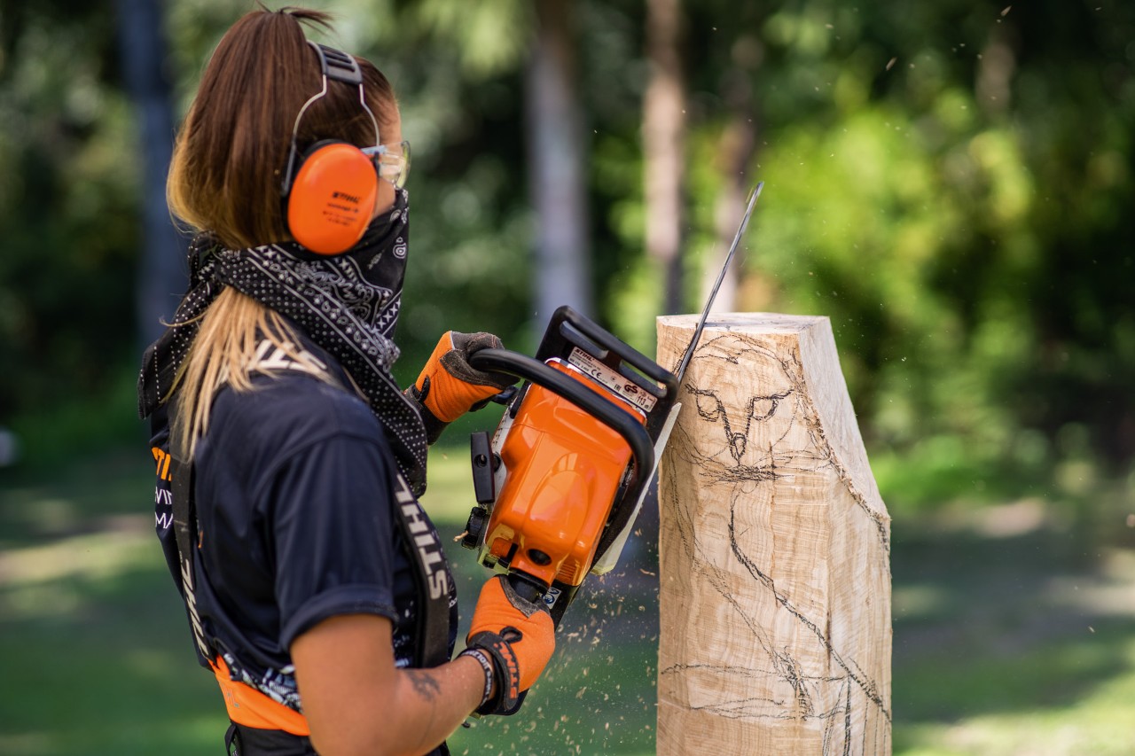 Femme avec casque antibruit, gants, bandana sur la bouche, utilisant une tronçonneuse STIHL pour sculpter un hibou