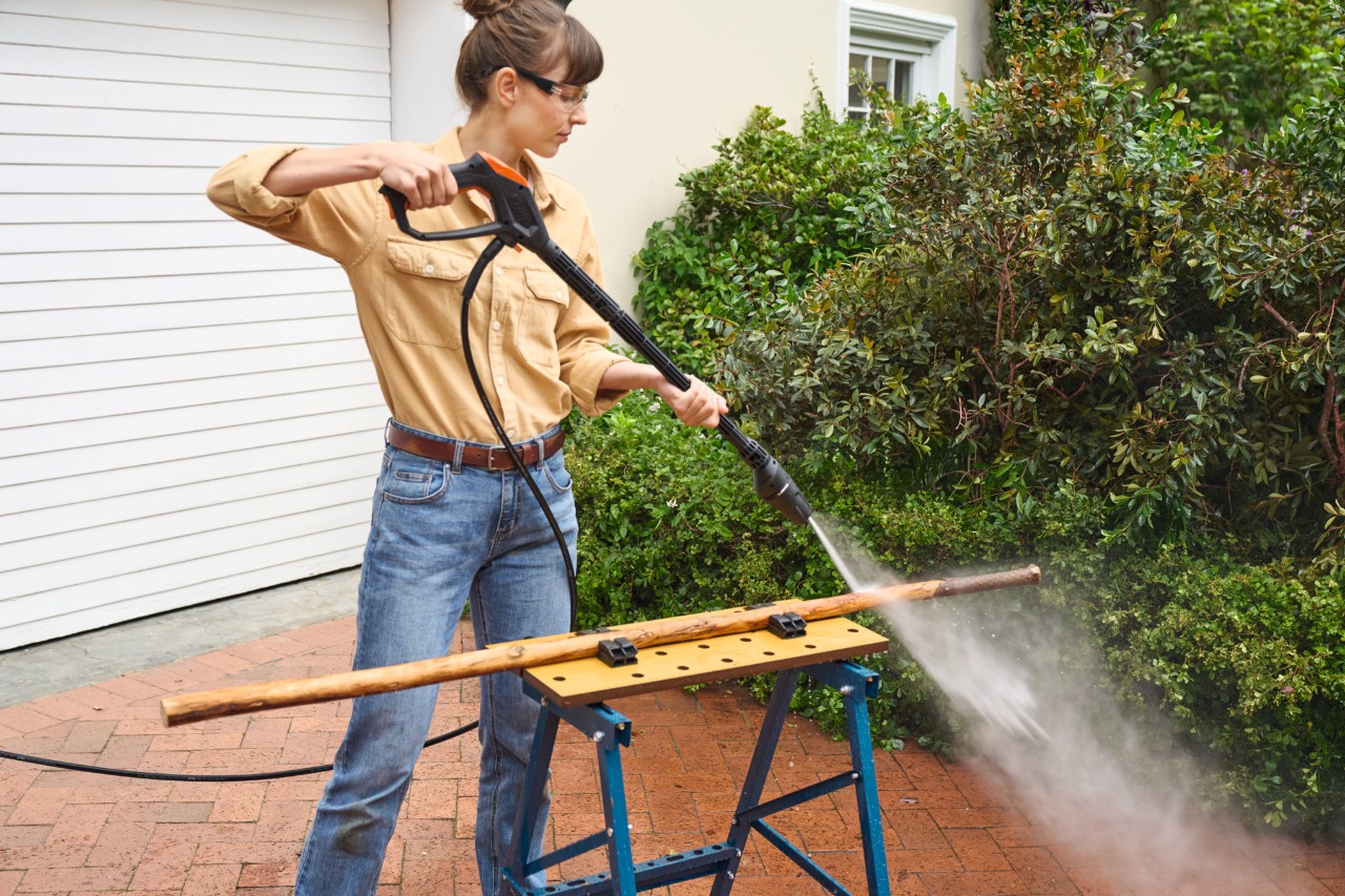 Dans une allée de garage, femme qui écorce une branche sur un établi à l’aide d’un nettoyeur haute pression STIHL RE 100
