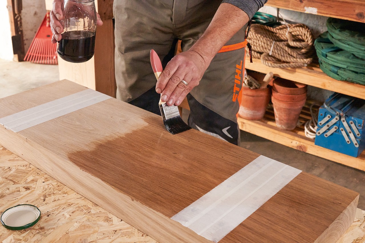 Un homme portant un pantalon anti-coupure STIHL huile une poutre en bois avec une brosse