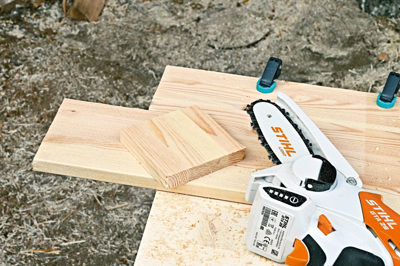 Une scie de jardin STIHL GTA 26 à côté d’une planche de bois dont un carré a été découpé 