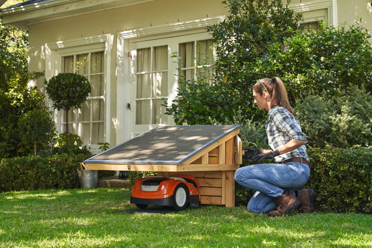 Femme sur un terrain de jardin avec un garage pour robot de tonte qu'elle a construit elle-même, y compris un robot de tonte STIHL, avec une maison en arrière-plan. 