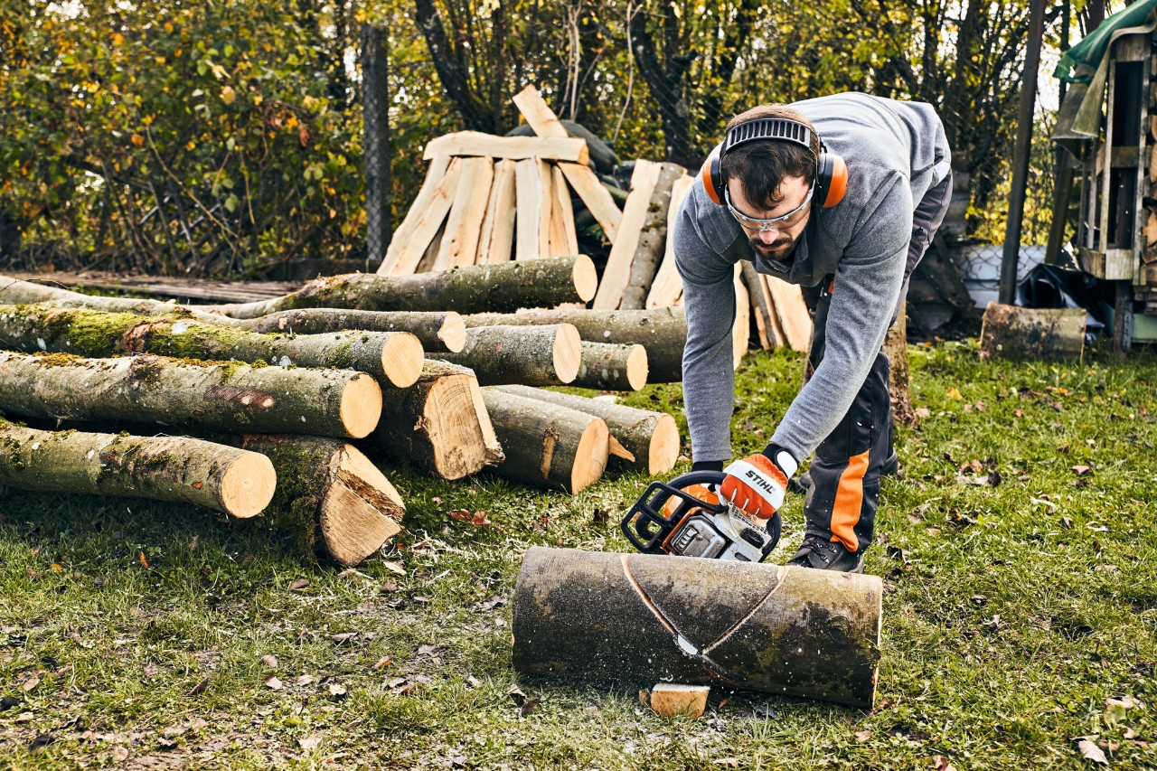 Un homme portant un équipement de protection du travail scie un tronc d'arbre pour construire lui-même un banc de jardin. 