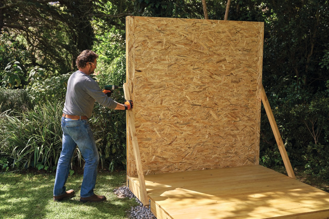 Un homme utilise des supports temporaires pour monter une paroi pré-assemblée sur la base d’un abri de jardin en bois