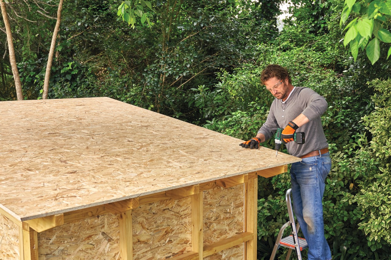 Comment construire un abri de jardin : homme sur une échelle vissant le panneau de toit en OSB sur un abri de jardin