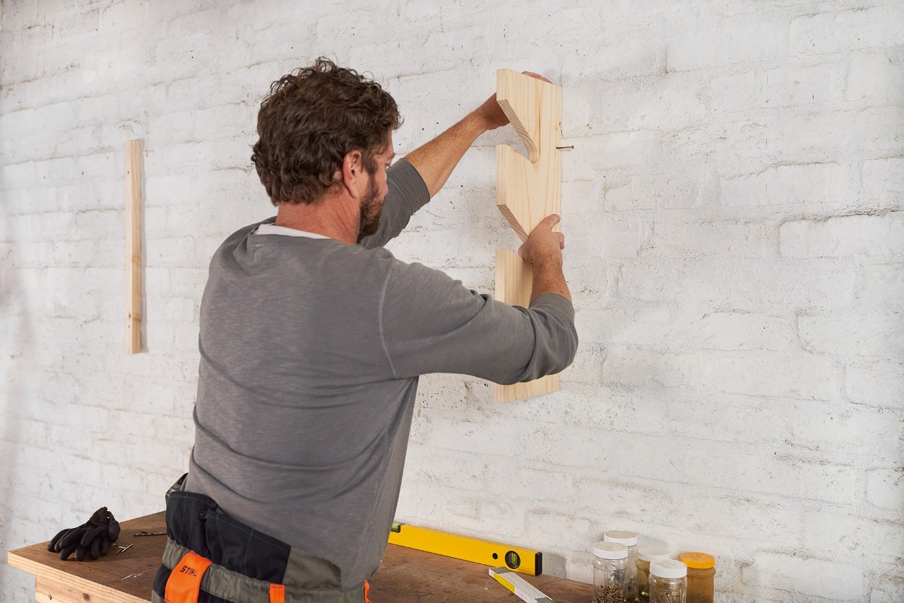 Un homme cheville une bande de bois au mur pour fabriquer lui-même un support de rangement pour outils de jardin 
