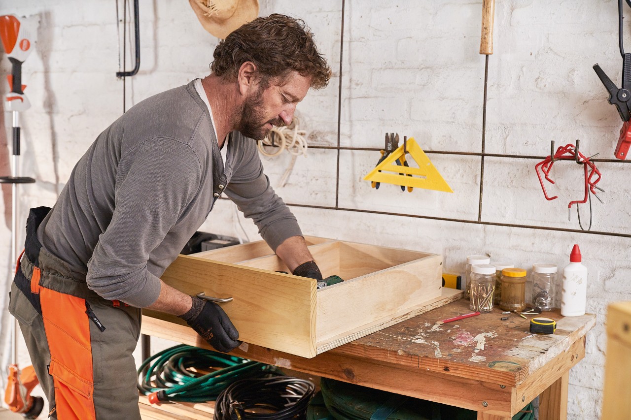 Un homme fixe une planche de pin à un tiroir pour fabriquer lui-même un rangement pour tondeuse à gazon