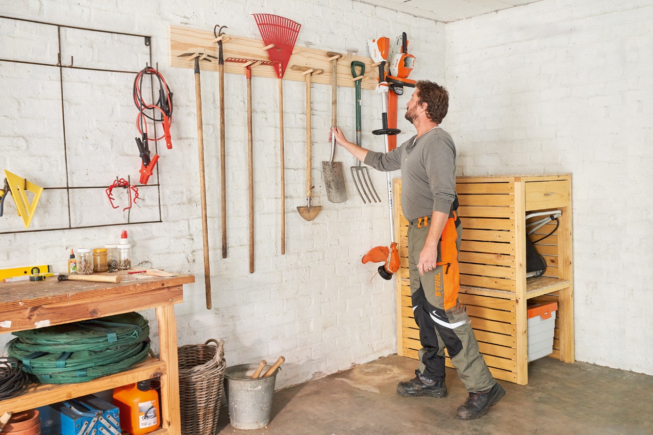 Homme en pantalon de travail STIHL accroche une pelle à un support pour outils  à côté d’un coupe-bordure STIHL 