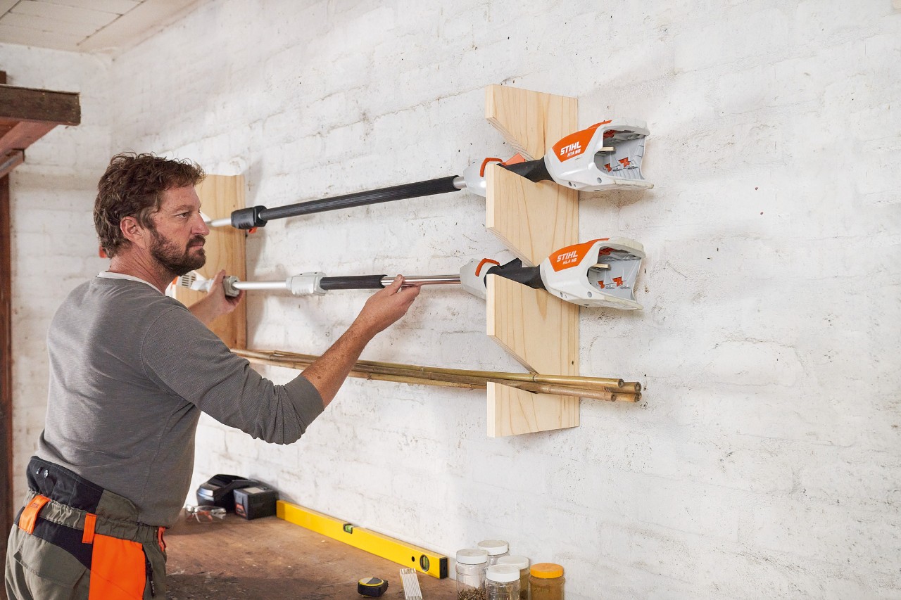 Homme place une débroussailleuse STIHL dans un support pour outils mural qu’il a fabriqué lui-même et fixé au mur 