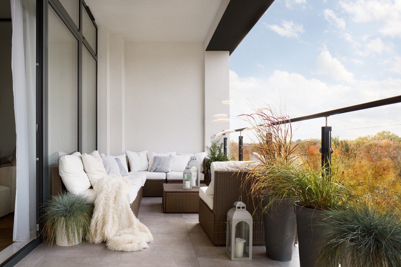 Des pots de graminées ornementales comme brise-vue sur un balcon, derrière un coin salon avec des coussins blancs ainsi que des lanternes  