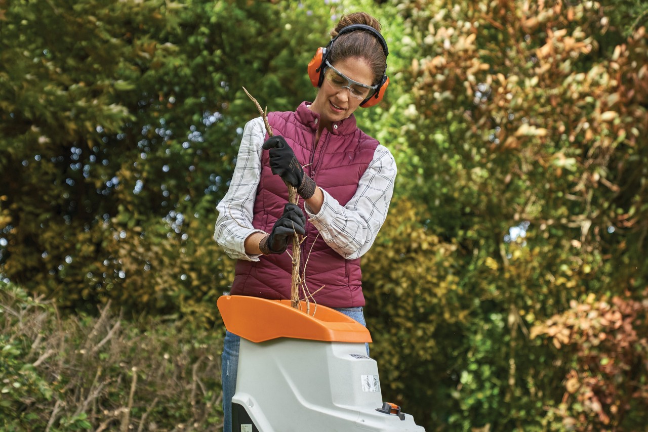 Femme portant des gants, des protections auditives et des lunettes insérant des branches dans un broyeur de jardin