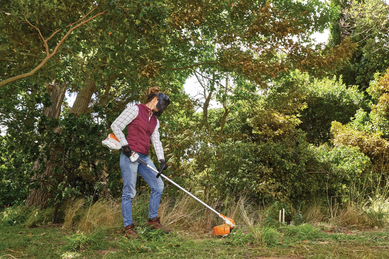 Femme utilisant une débroussailleuse sur des hautes herbes avec des arbres et des arbustes en arrière-plan
