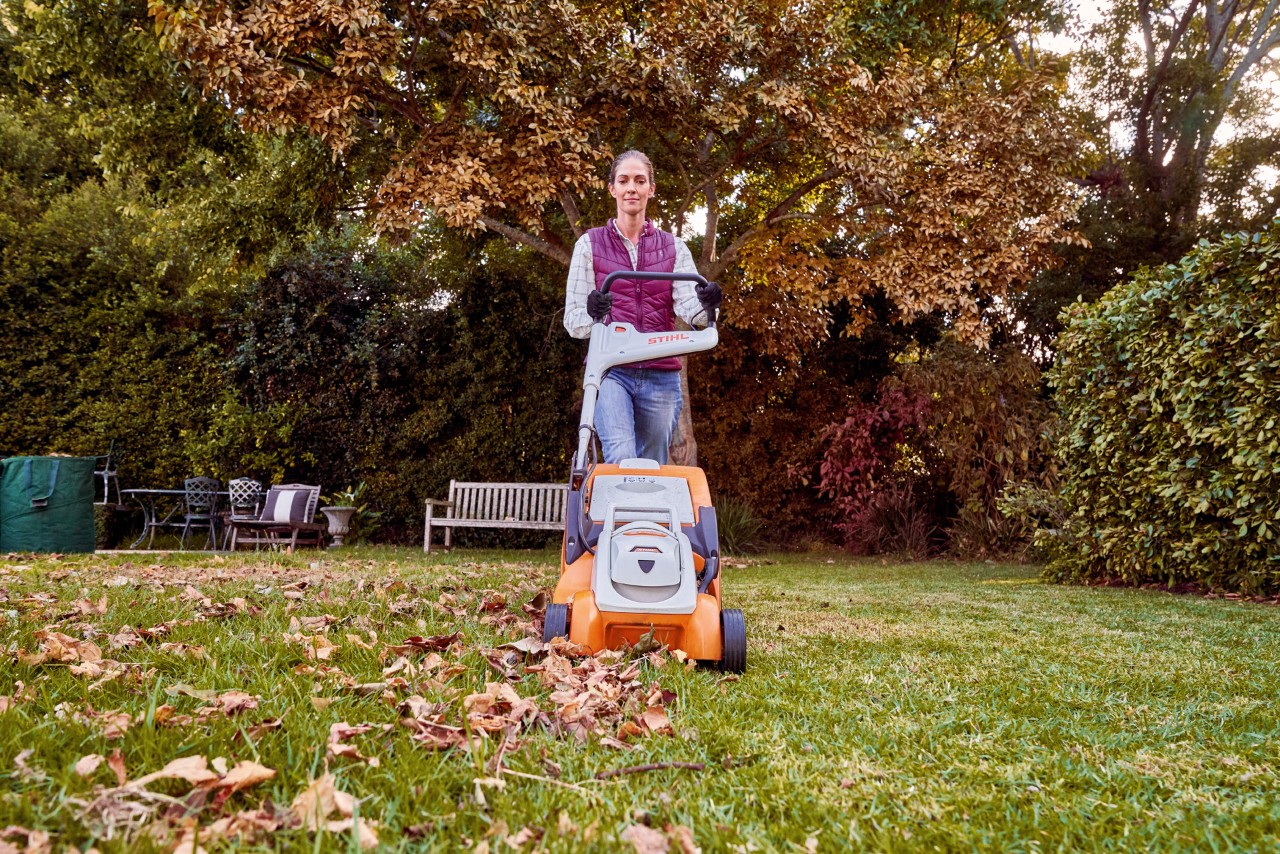 Femme tondant une pelouse de jardin recouverte de feuilles à l’automne avec la tondeuse à gazon sans fil STIHL RMA 339 C
