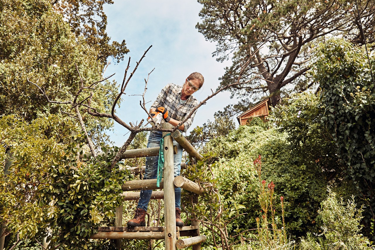 Une femme installée sur un support en bois dans un jardin, qui coupe une branche avec une scie de jardin à batterie STIHL GTA 26
