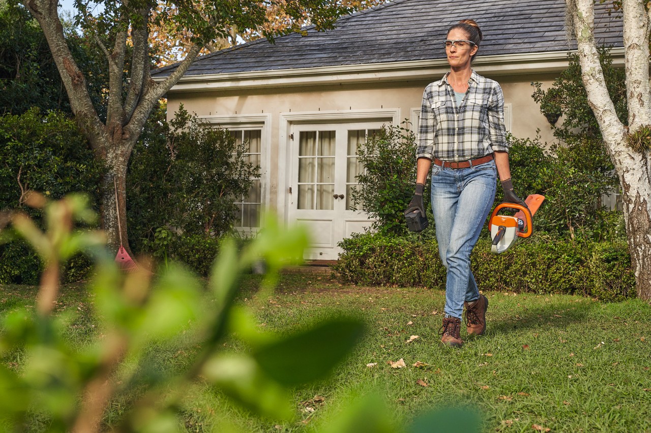 Une femme marche avec un taille-haies STIHL sur la pelouse devant une maison