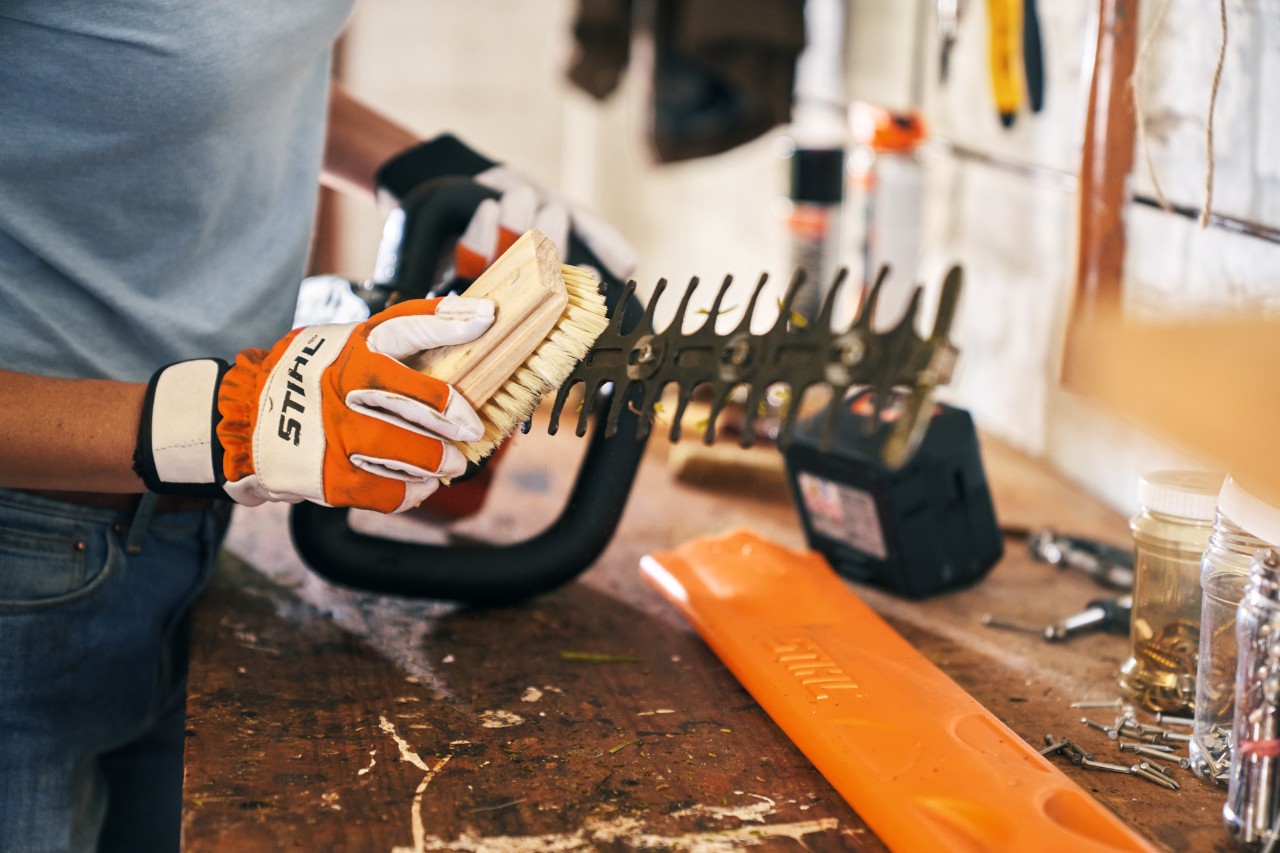Une personne portant des gants de sécurité nettoie un taille-haie à l’aide d’une brosse dans un atelier 