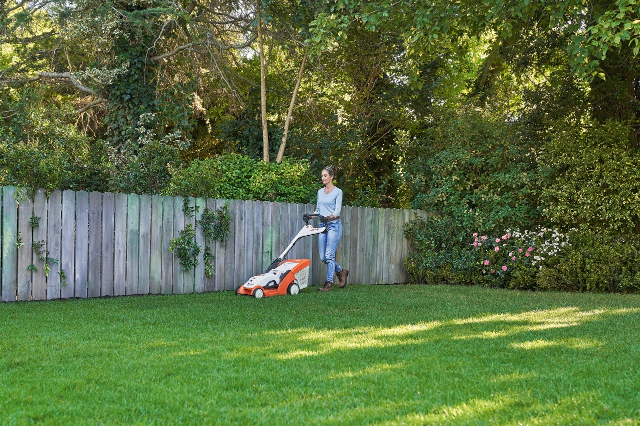 Une femme tond une pelouse verte devant une clôture en bois avec la tondeuse à gazon à batterie STIHL RMA 339 C.