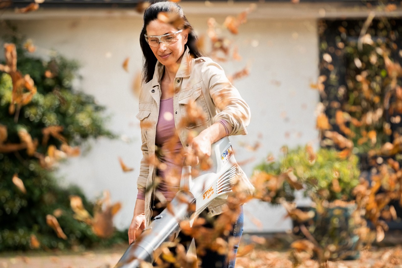 Une femme nettoie sa pelouse au printemps avec un souffleur sur batterie STIHL BGA 56 alors que des feuilles s’envolent