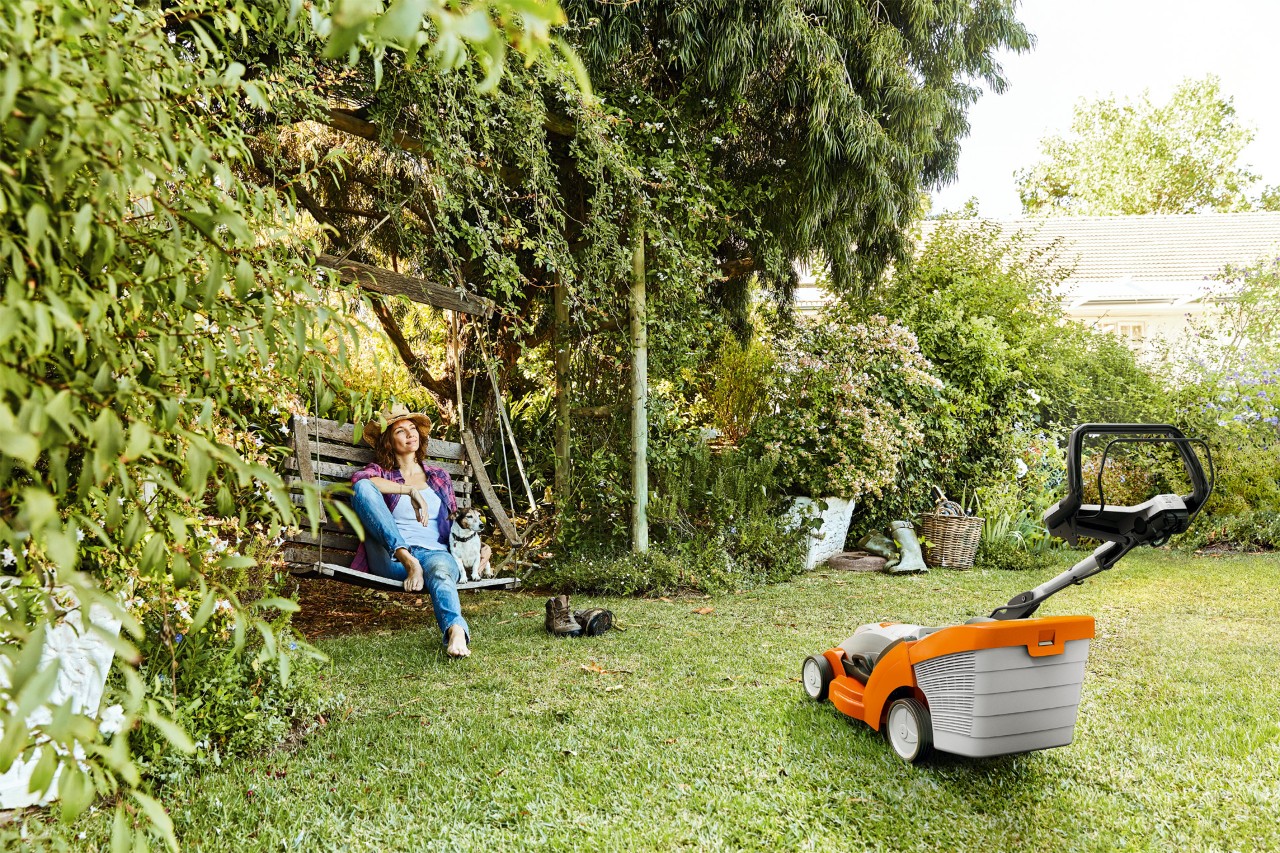Femme détendue sur une balançoire de jardin à côté d’une tondeuse à gazon à batterie STIHL RMA 339 sur un terrain gazonné