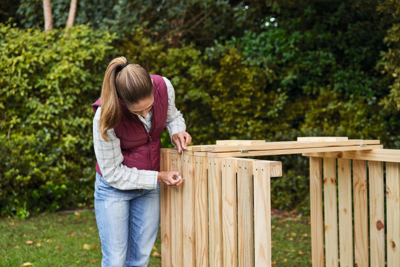 Une femme dans un jardin fixe des crochets à vent au bac à compost qu’elle a construit elle-même 