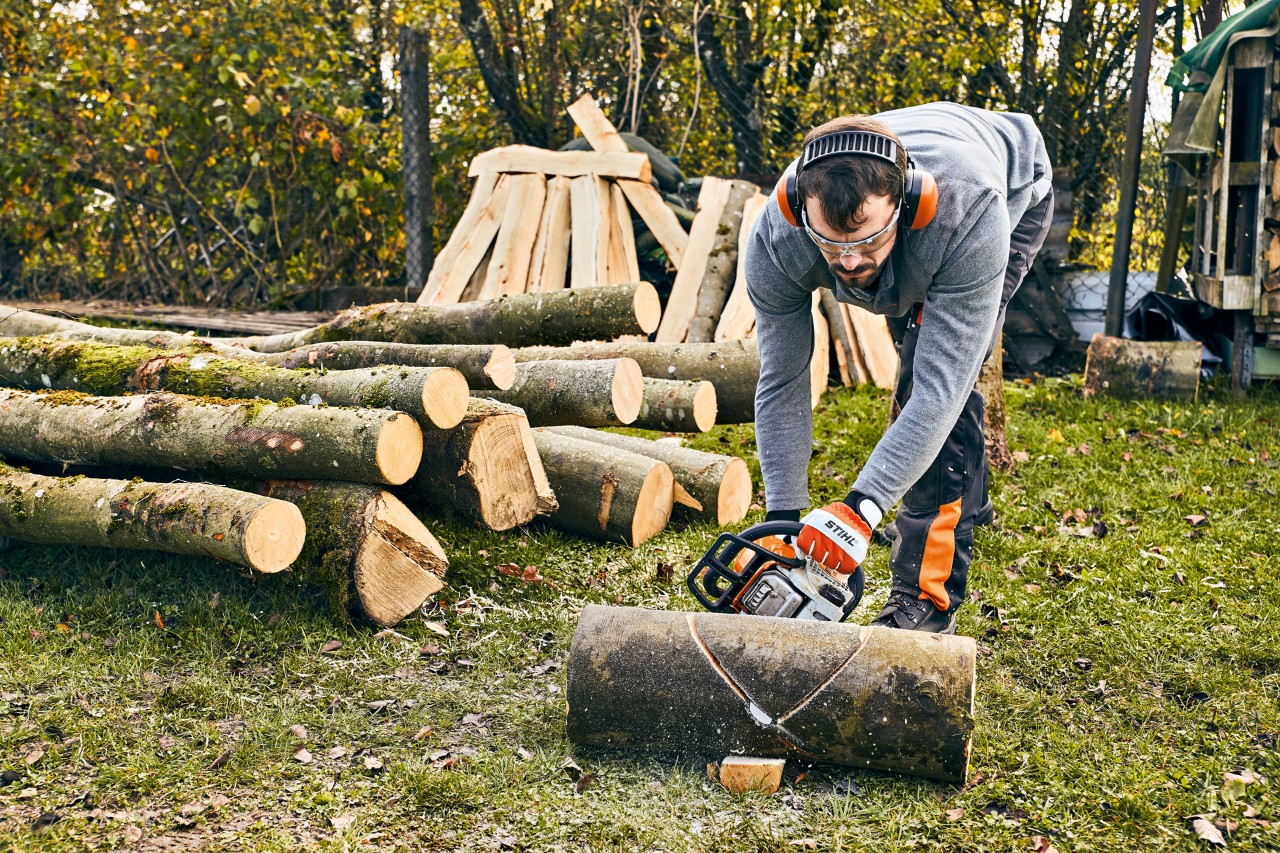 Homme devant une pile de bois, vêtu d’une tenue de protection fait une entaille en V dans un tronc avec une tronçonneuse
