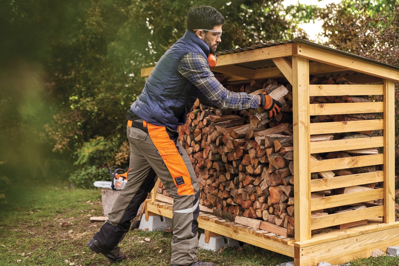Un homme en vêtements de protection empile du bois de chauffage dans un abri à bois de chauffage diy