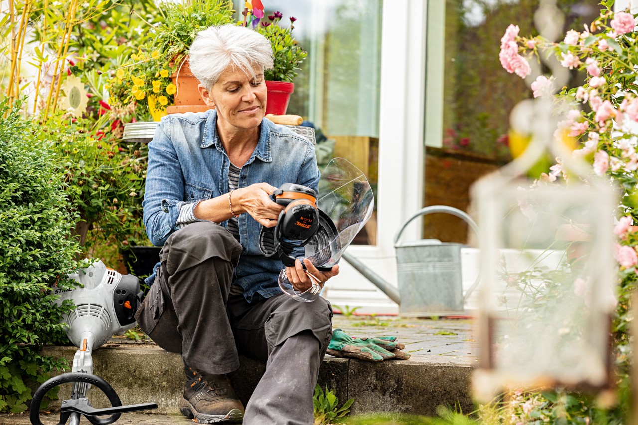 Femme assise entre des plantes en fleurs à côté d’une débroussailleuse STIHL FS&nbsp;38 et examine l’équipement de protection