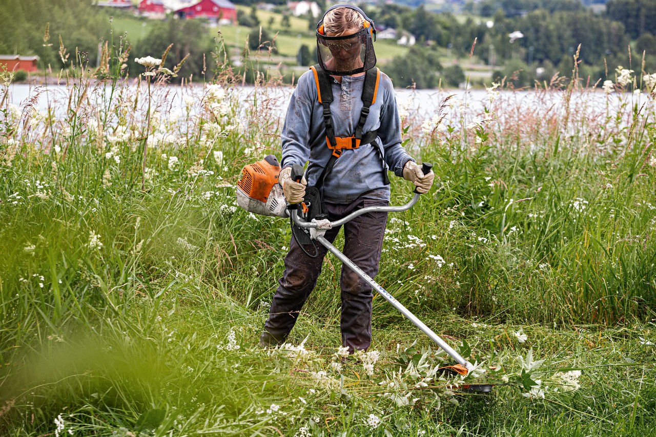 Une femme équipée d’une visière utilisant une débroussailleuse thermique STIHL FS 240 sur de l’herbe haute près d’un lac
