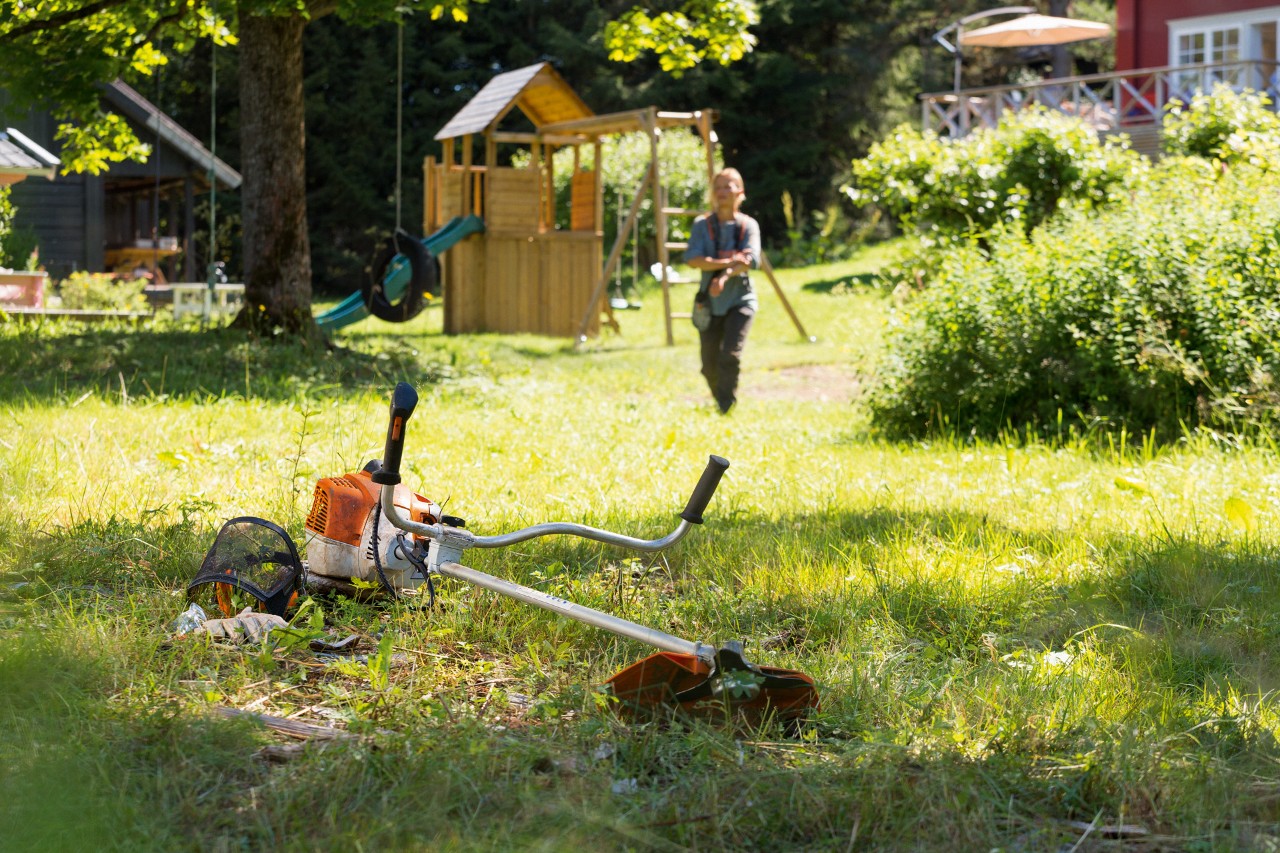 Débroussailleuse STIHL FS 240 au sol dans un jardin, avec des jeux pour enfant et un homme s’approchant de l’outil