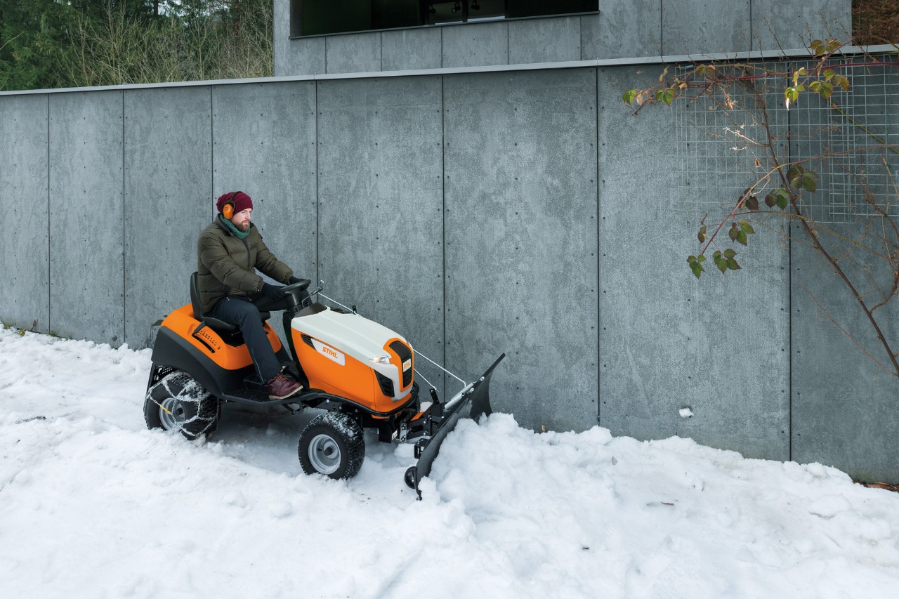 Un homme déblaie la neige devant un mur sur un tracteur tondeuse équipé d’une lame chasse-neige 