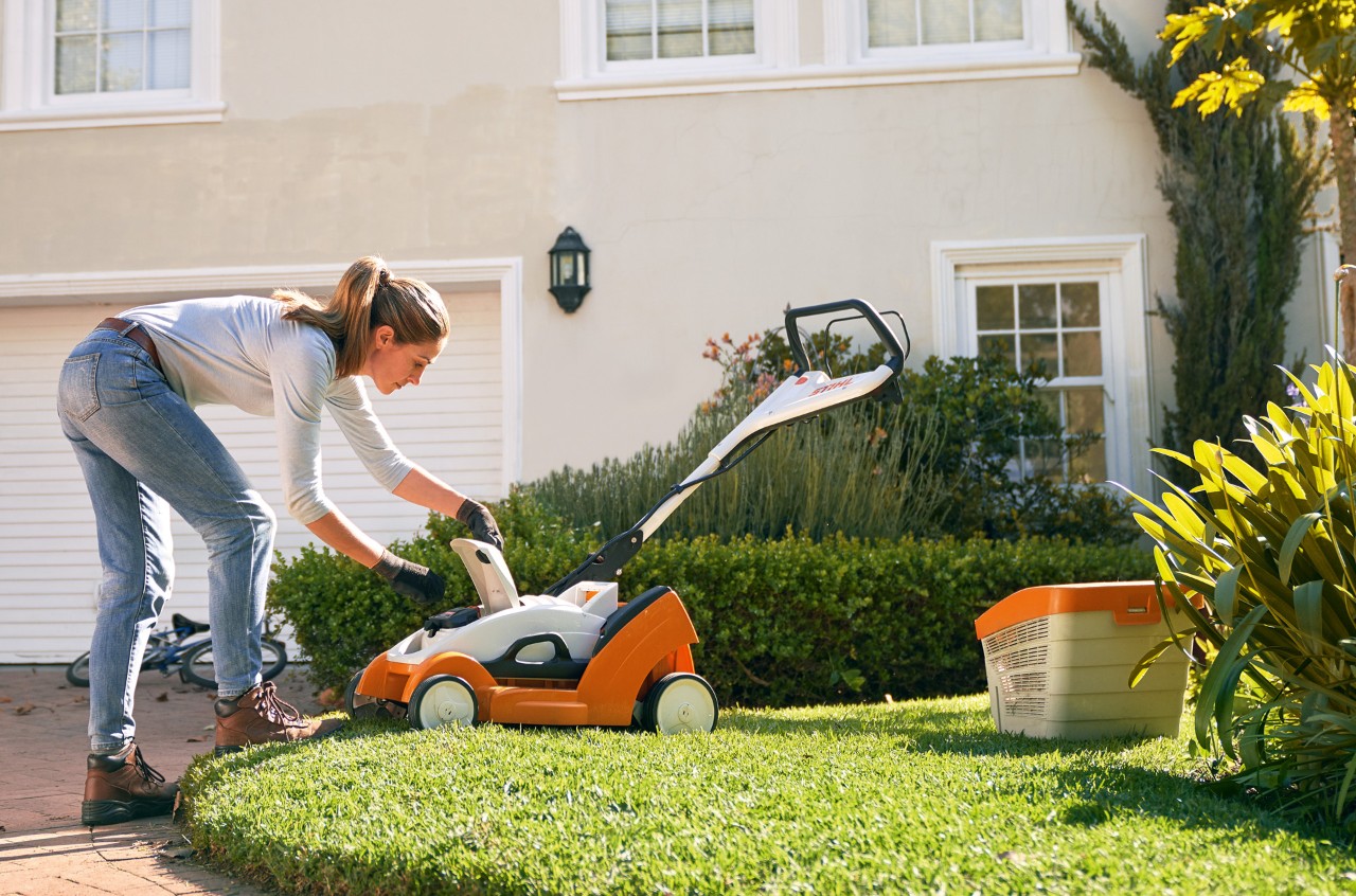Femme ouvrant le couvercle d’une tondeuse à gazon sur batterie STIHL RMA 339 C devant une maison blanche