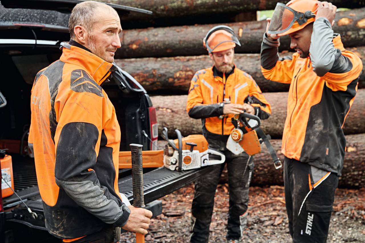 Trois hommes en équipement de protection STIHL avec des outils à batterie STIHL à côté du coffre ouvert d’un véhicule, en arrière-plan un tas de bois