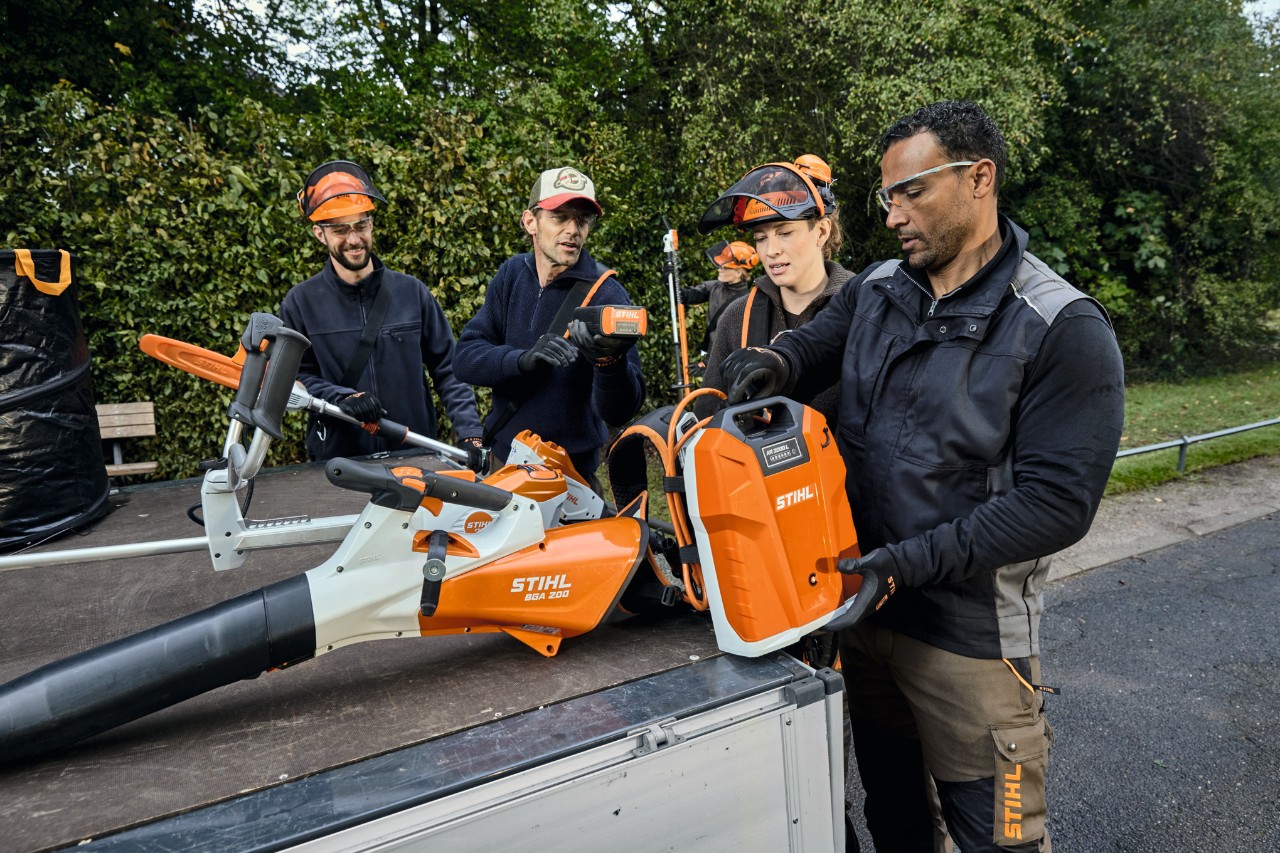 Trois hommes et une femme en équipement de protection STIHL se tiennent près d’une surface de chargement d’un véhicule équipé d’appareils STIHL, par exemple le souffleur à batterie STIHL BGA 200 et la batterie dorsale STIHL AR 2000 L 