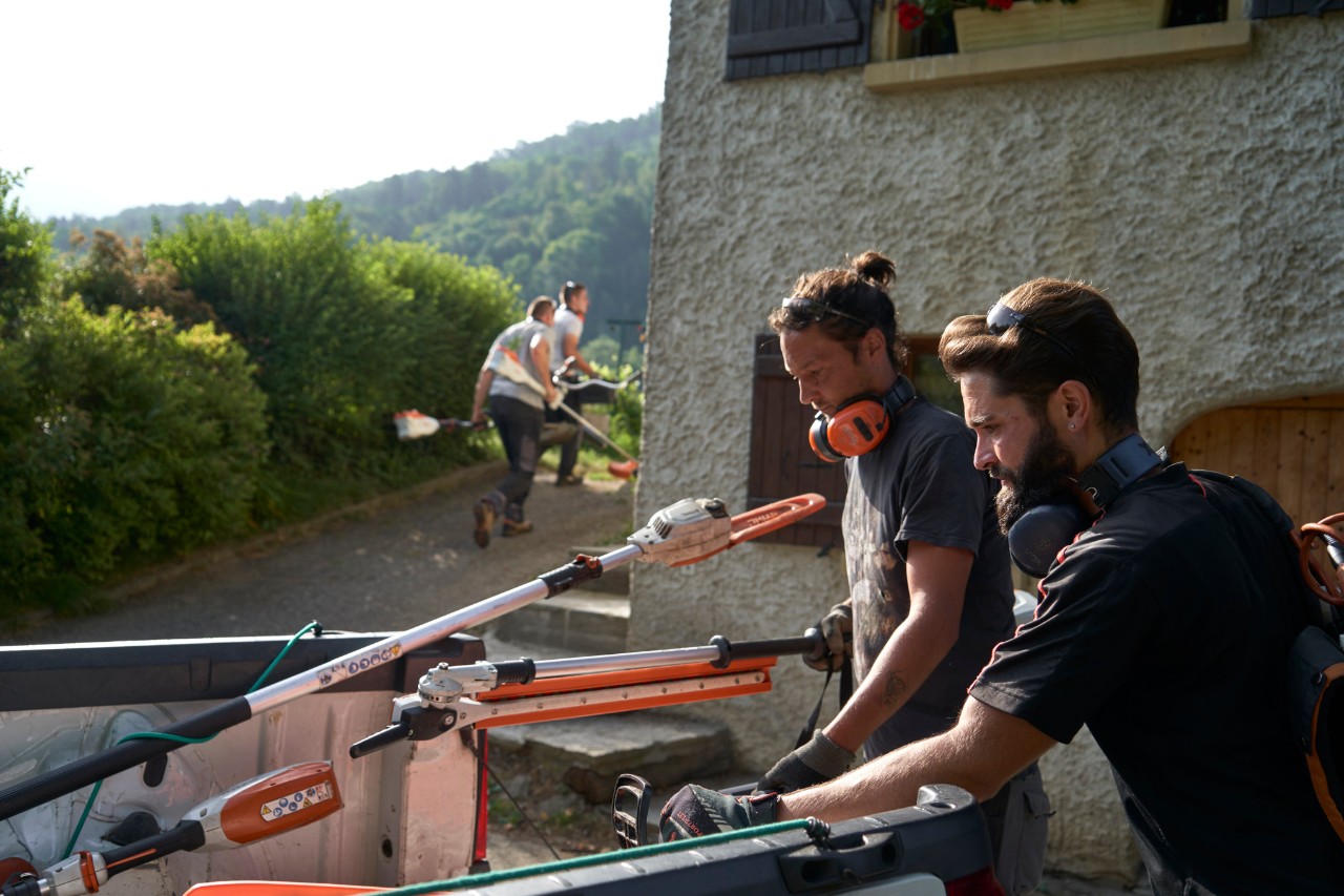 Deux paysagistes déchargent des outils à batterie STIHL d'une voiture, d'autres ouvriers sont à l'arrière-plan. 