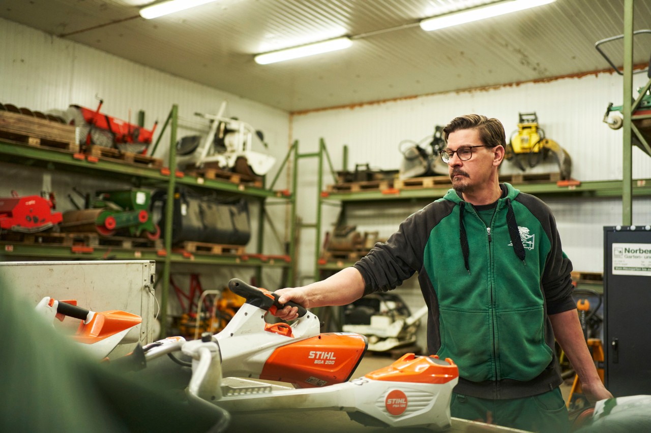 Homme dans un atelier se tenant derrière une surface de travail avec des appareils à batterie STIHL, attrappant le souffleur à batterie STIHL BGA 200