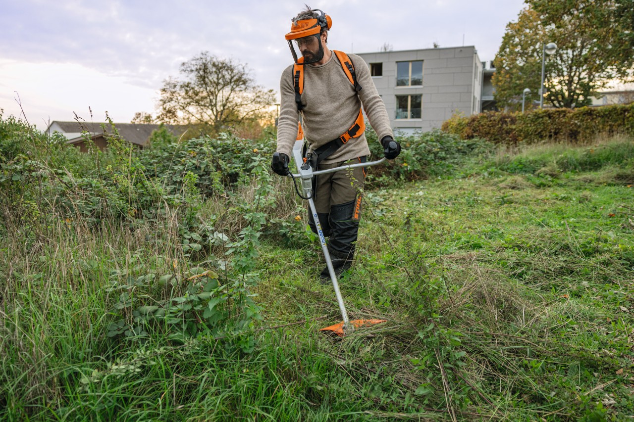 Homme en équipement de protection STIHL tondant un espace vert envahi par les mauvaises herbes avec une débroussailleuse à batterie STIHL FSA 200