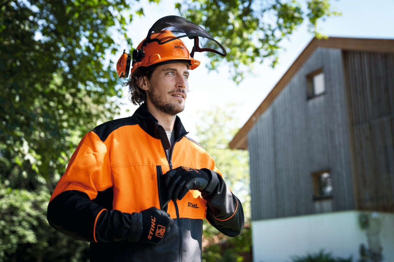 Homme portant un équipement de protection STIHL orange et noir, avec un casque et des gants, l’air confiant, devant une maison et des arbres en arrière-plan 