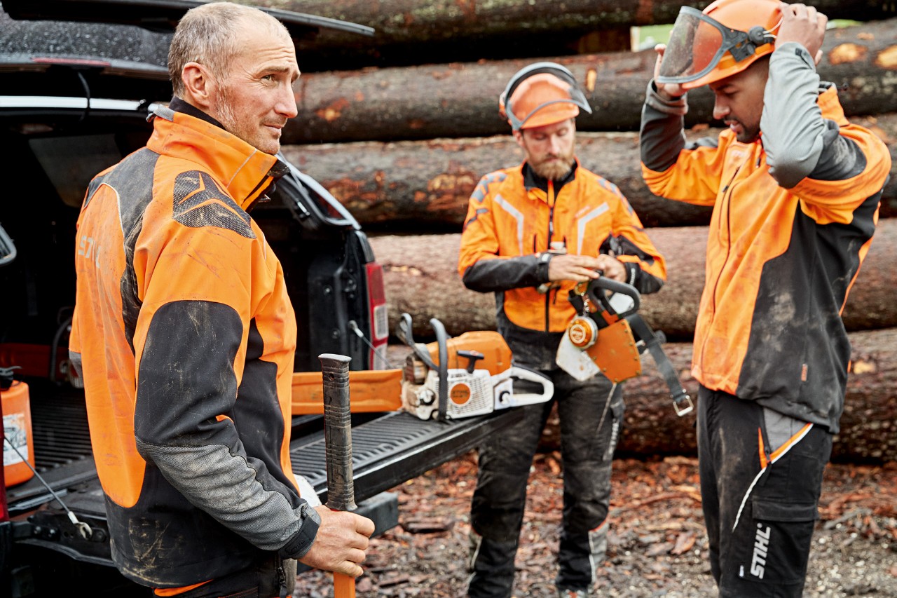 Trois hommes en vêtements de protection STIHL devant un tas de bois se préparant à travailler avec une tronçonneuse STIHL