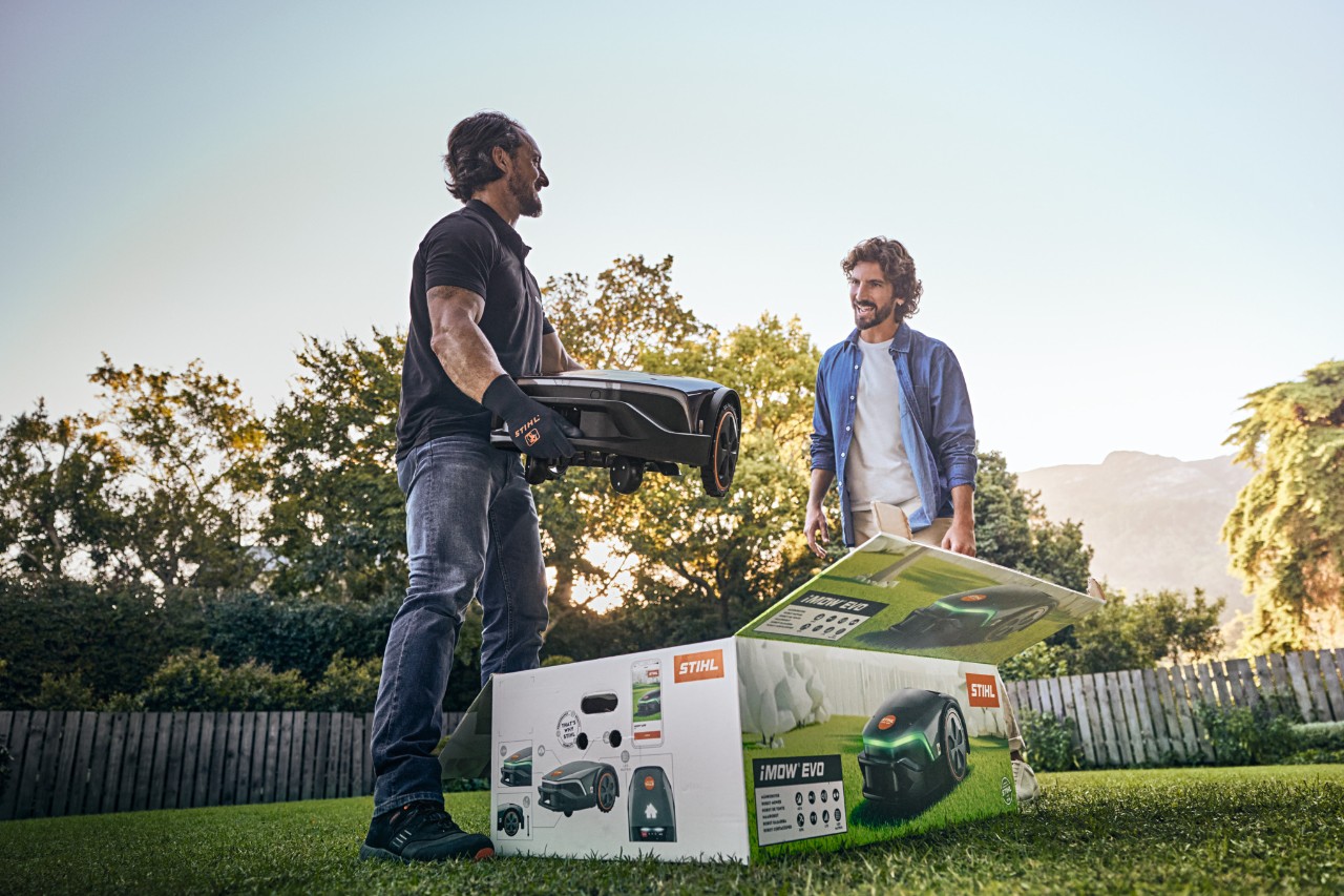 Carton iMOW® dans le jardin d’un client. Un revendeur STIHL porte une iMOW® EVO et souhaite aider à l’installer